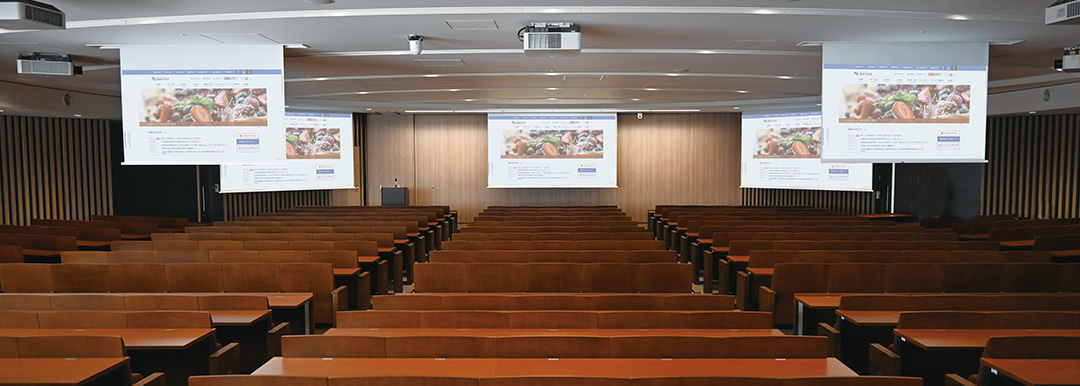 米田吉盛記念講堂<br>
					円形の講堂で、前面に150インチスクリーンを3台、後方には80インチスクリーンを2台設置し、どの席に座っても資料が確認できる
